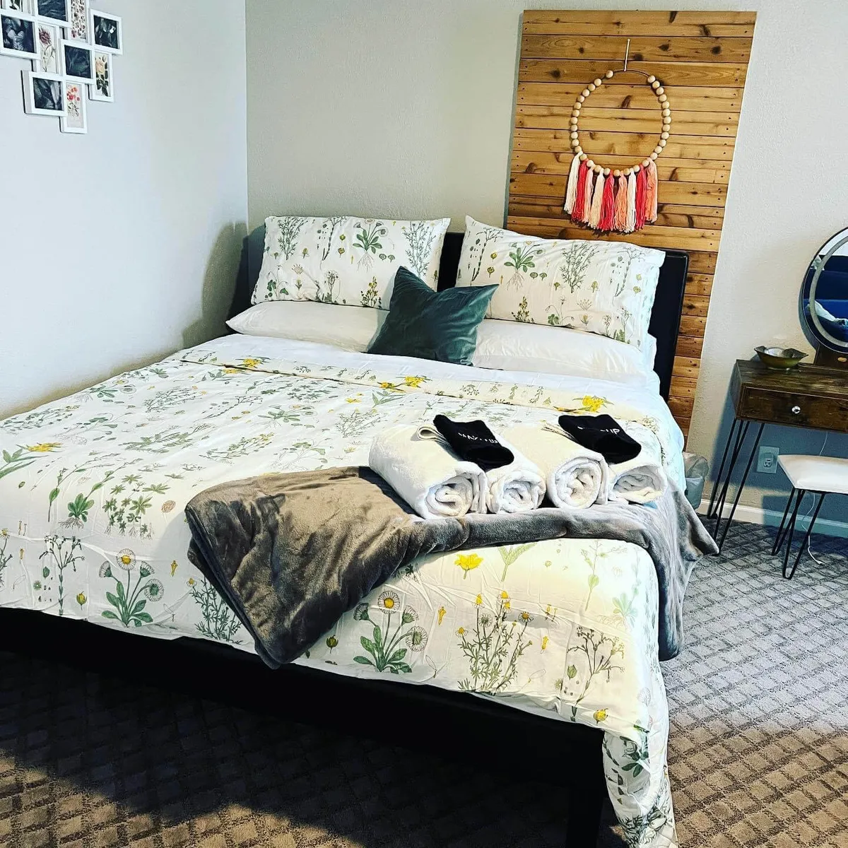 A bedroom scene featuring a bed with floral bedding set against a wooden accent wall. A decorative wall hanging and a mirrored bedside table add style and elegance to the private sleeping area. The soft, neutral color scheme contributes to the tranquil, restful mood.