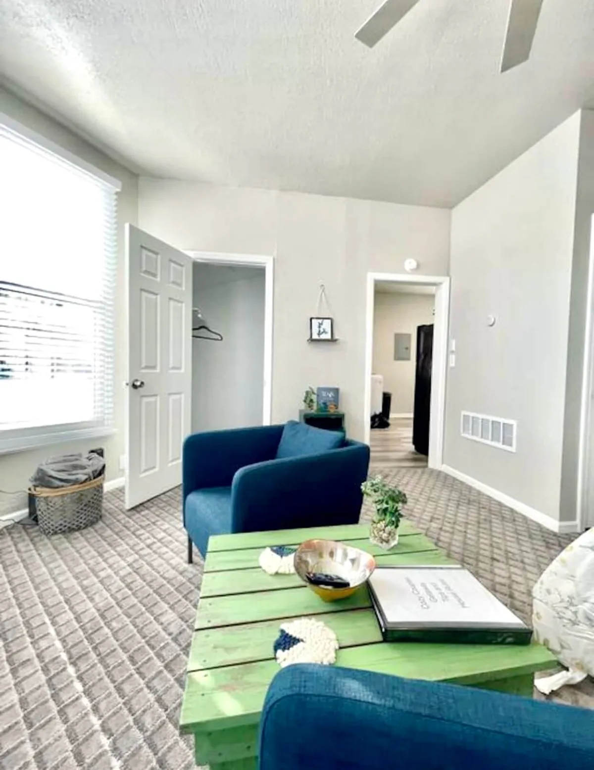 This image offers a close-in, intimate look at the lounge, focusing on the green coffee table and blue chairs beside a window with blinds. The natural lighting creates a warm, welcoming ambiance that invites moments of quiet reflection or shared stories over coffee.