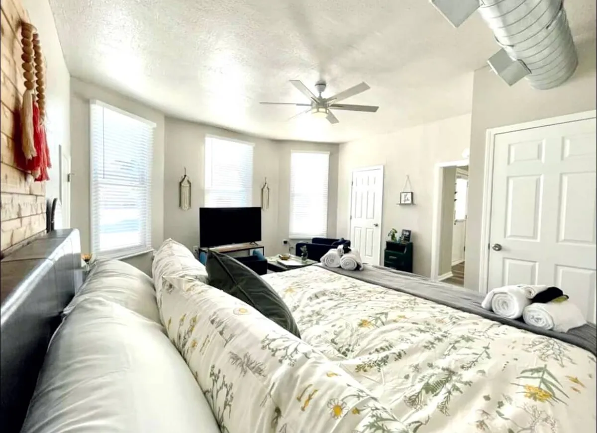 A spacious bedroom featuring a large bed with floral bedding as its centerpiece. The room is brightened by natural light streaming through windows, and a television is positioned for comfortable viewing from the bed. The overall color palette is soft and inviting, with plush linens and subtle decorative touches that create a serene, restful environment.