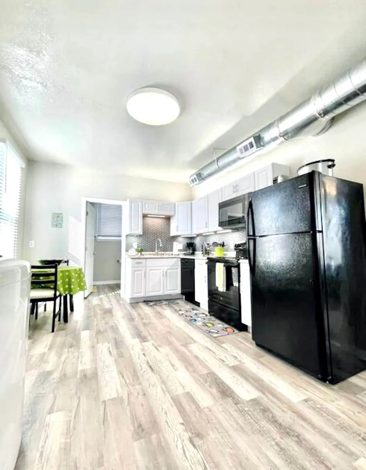 A wide-angle kitchen shot with modern appliances, white cabinetry, and a dining table set for a meal. The space feels warm and welcoming, ideal for both family meals and entertaining guests. The layout emphasizes openness and practicality, with everything easily accessible.