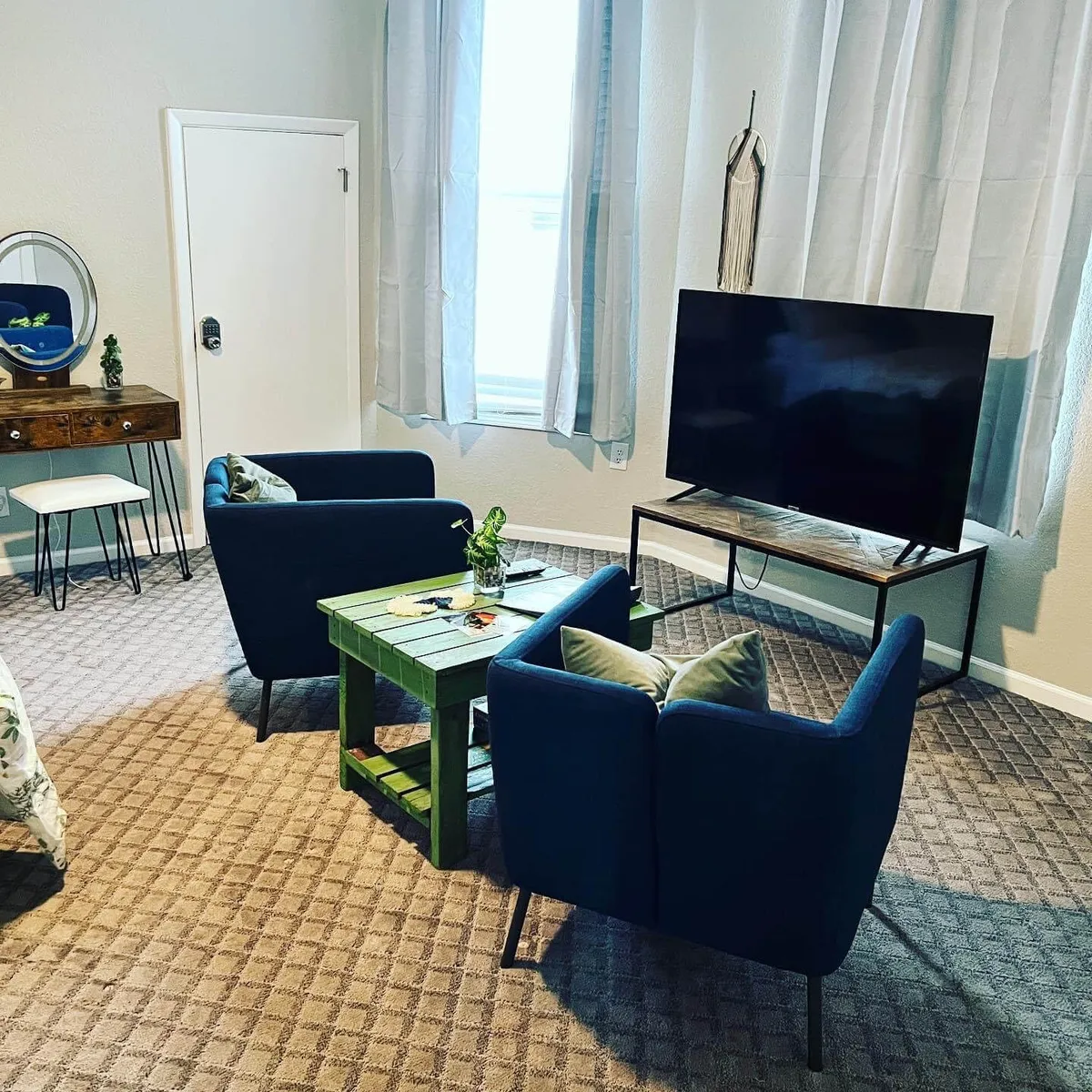 A slightly different angle of the lounge area, this image features a TV, the green coffee table, blue chairs, and a mirror with a plant. The arrangement creates a sense of homey intimacy and offers additional context for the space's versatility—ideal for watching TV, reading, or chatting over coffee.