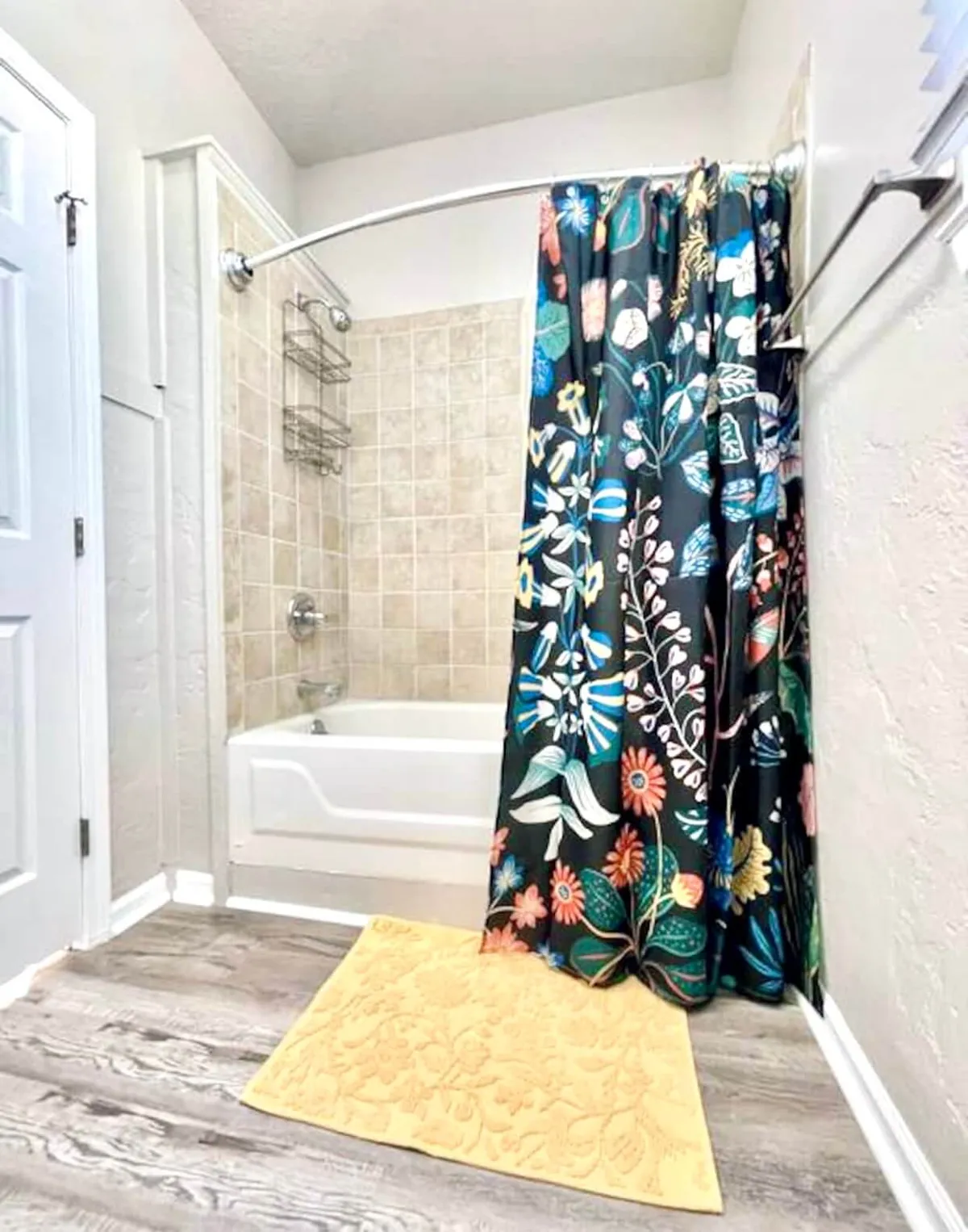 A cheerful bathroom scene showcasing a floral shower curtain, a soft yellow bath mat, and a clean bathtub. The combination of fresh textiles and sparkling surfaces exudes warmth and comfort, perfect for self-care routines.