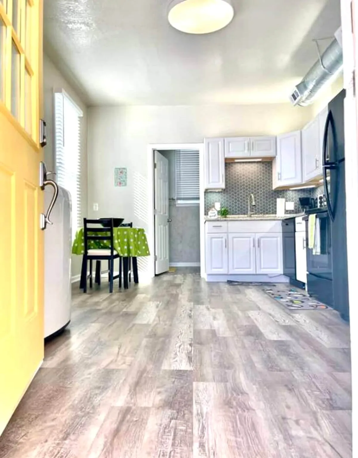 A cheerful kitchen scene featuring a yellow door that adds a pop of color, a green polka dot tablecloth over a dining table, and bright white cabinetry. Modern kitchen appliances are visible, and the layout feels open and welcoming. The space invites both cooking and casual dining, with plenty of natural light enhancing the atmosphere.