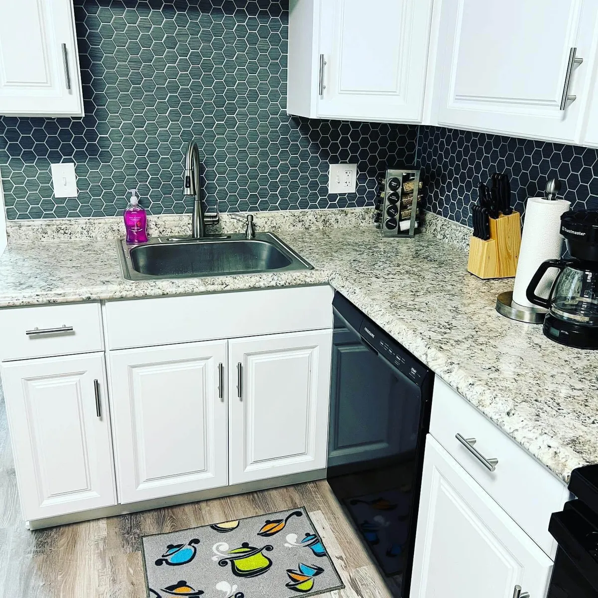 A close-up of a remodeled kitchen workspace showcasing a gleaming granite countertop, a stylish hexagonal backsplash, a sink area, and a coffee maker. A colorful mat adds vibrancy to the practical space. The area is tidy and modern, emphasizing updated finishes and functionality.