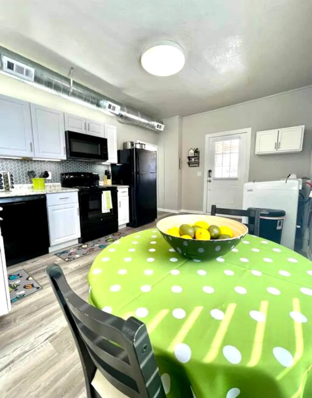 This image features a kitchen or dining area with a cheerful green tablecloth adorned with polka dots, a bowl of fresh fruit, and visible black appliances. The setting is bright, inviting, and designed for gathering, with elements suggesting meal preparation or enjoying food together.