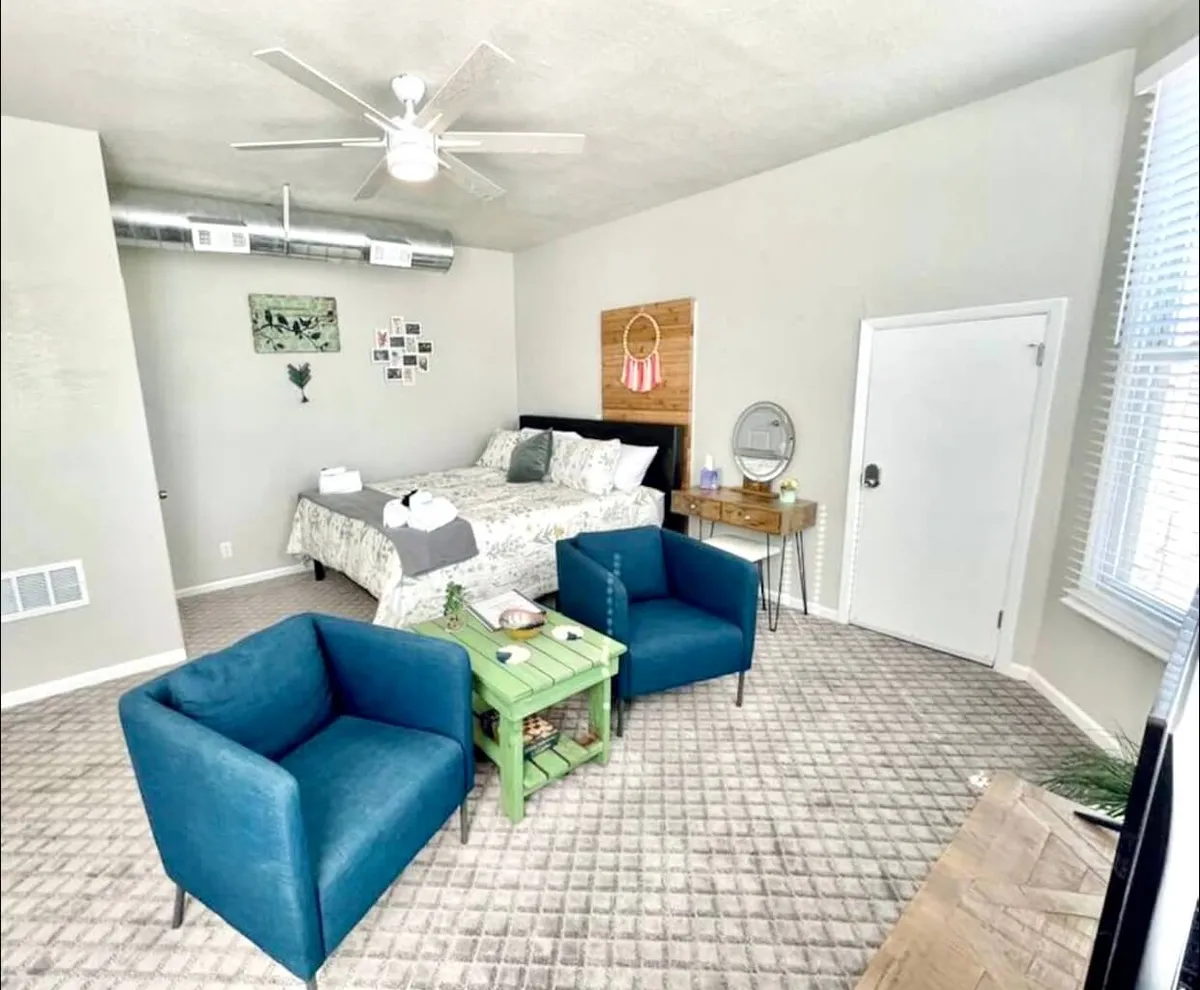 This image captures the inviting bedroom space with a California King bed adorned in floral bedding. A green accent table and blue chairs add vibrant touches, and the room exudes a feeling of comfort and relaxation—a place to unwind after dining out or exploring the local area.