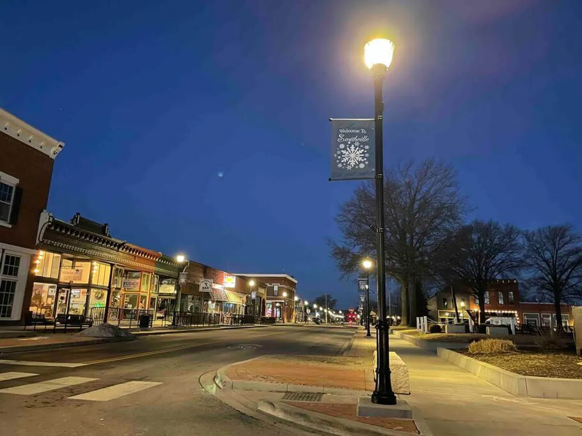 The image captures Smithville’s historic downtown, featuring warmly lit street lamps, charming brick facades, and a festive welcome banner strung across the avenue. The scene is alive with the atmosphere of evening strolls, visible boutique storefronts, and hints of local dining establishments, giving a sense of the vibrant community.