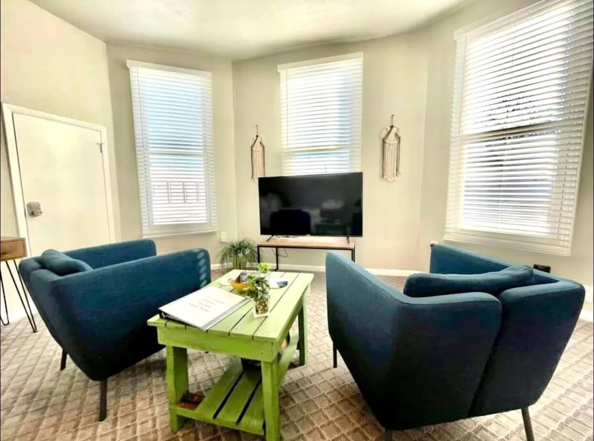 This image shows a cozy living area with blue armchairs arranged around a green coffee table. A television is visible, and natural light filters in through window treatments, casting a warm glow over the space. The setup suggests comfort and encourages both socializing and relaxation.