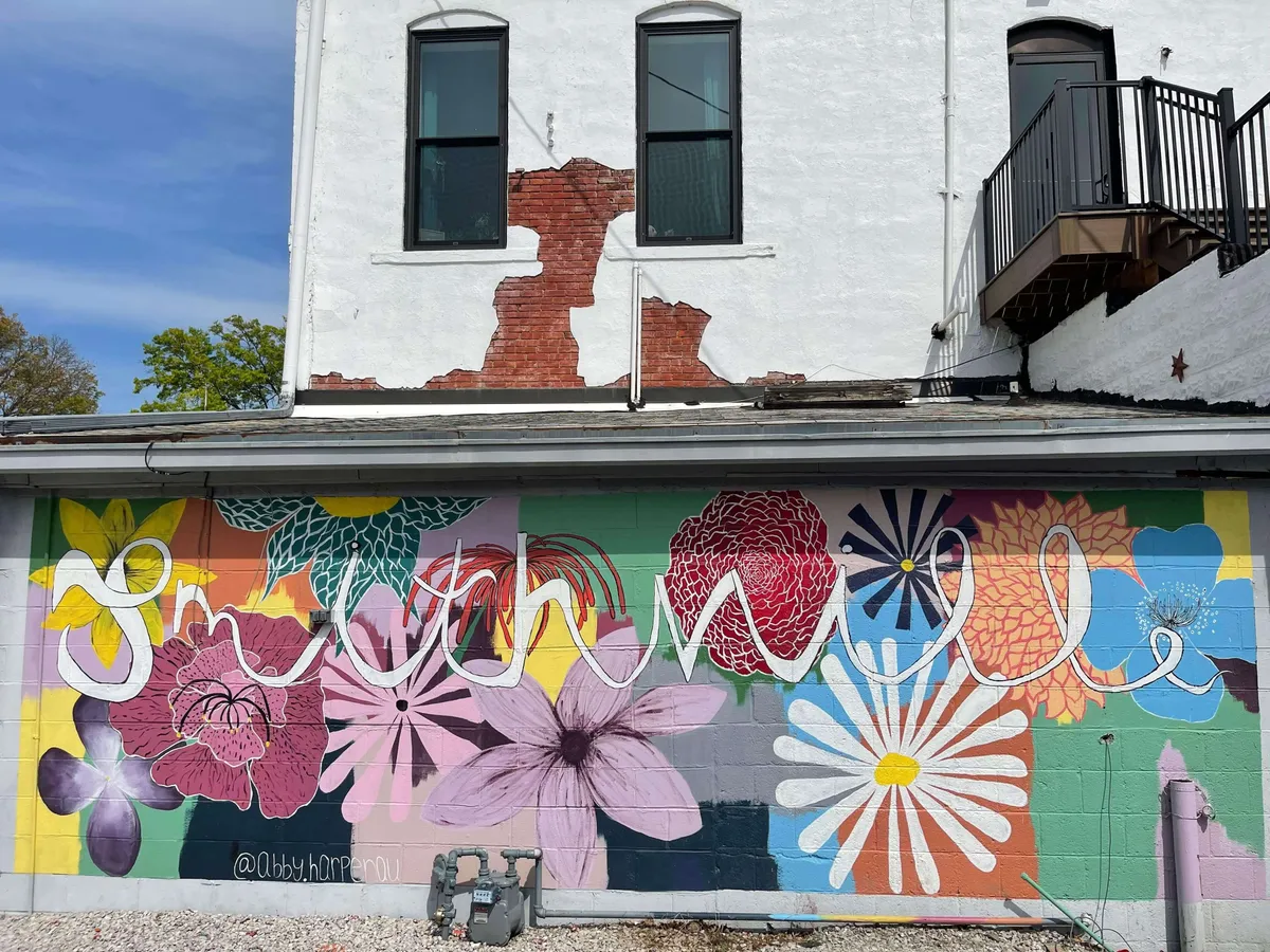 This exterior image features a colorful mural painted on a textured wall, with the word 'smooth' prominently displayed. Locals and visitors are seen admiring the artwork, capturing the playful spirit of Smithville’s arts scene and its unique community character.