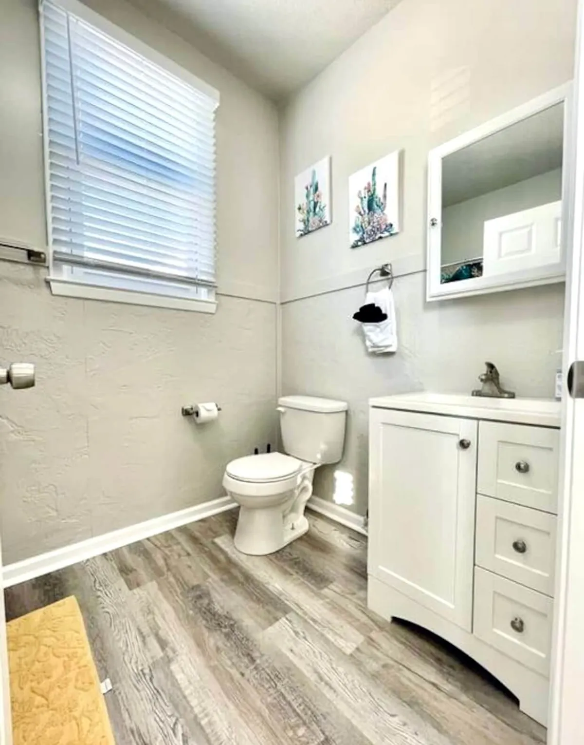 A bright and airy bathroom featuring a modern vanity with sink, complemented by subtle artwork and a window with blinds that filters natural light. The uncluttered surfaces emphasize functionality and comfort.